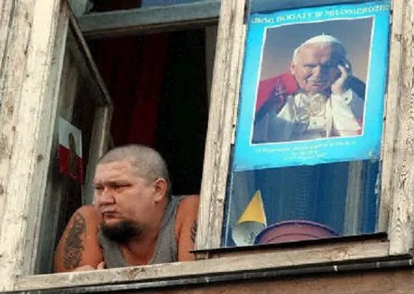 Mężczyzna z tatuażami wygląda przez okno starej kamienicy, obok w szybie wisi plakat z Janem Pawłem II i napisem „Bóg bogaty w miłosierdzie”