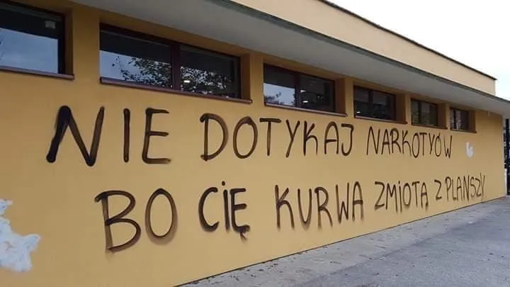 Żółta ściana budynku z odręcznym czarnym napisem ostrzegającym przed narkotykami, zakończonym słowami „zmiotą z planszy”