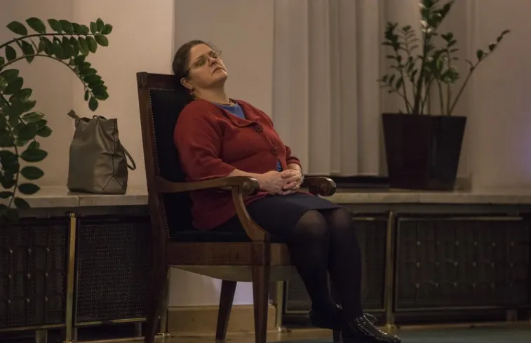 Krystyna Pawłowicz w czerwonym swetrze siedzi na drewnianym krześle z odchyloną głową i zamkniętymi oczami