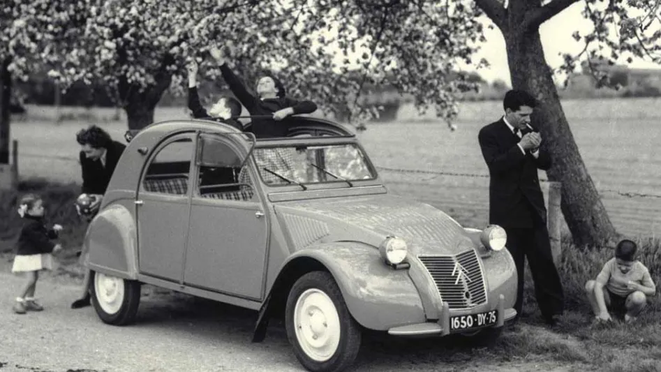 Szary Citroën 2CV z lat 50. zaparkowany pod kwitnącym drzewem; obok kobieta z dzieckiem, dwoje dzieci wychyla się przez otwarty dach, mężczyzna zapala papierosa